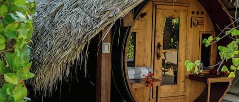 Una estructura de madera con techo de paja, una puerta circular y un pequeño porche con una silla y una mesa.