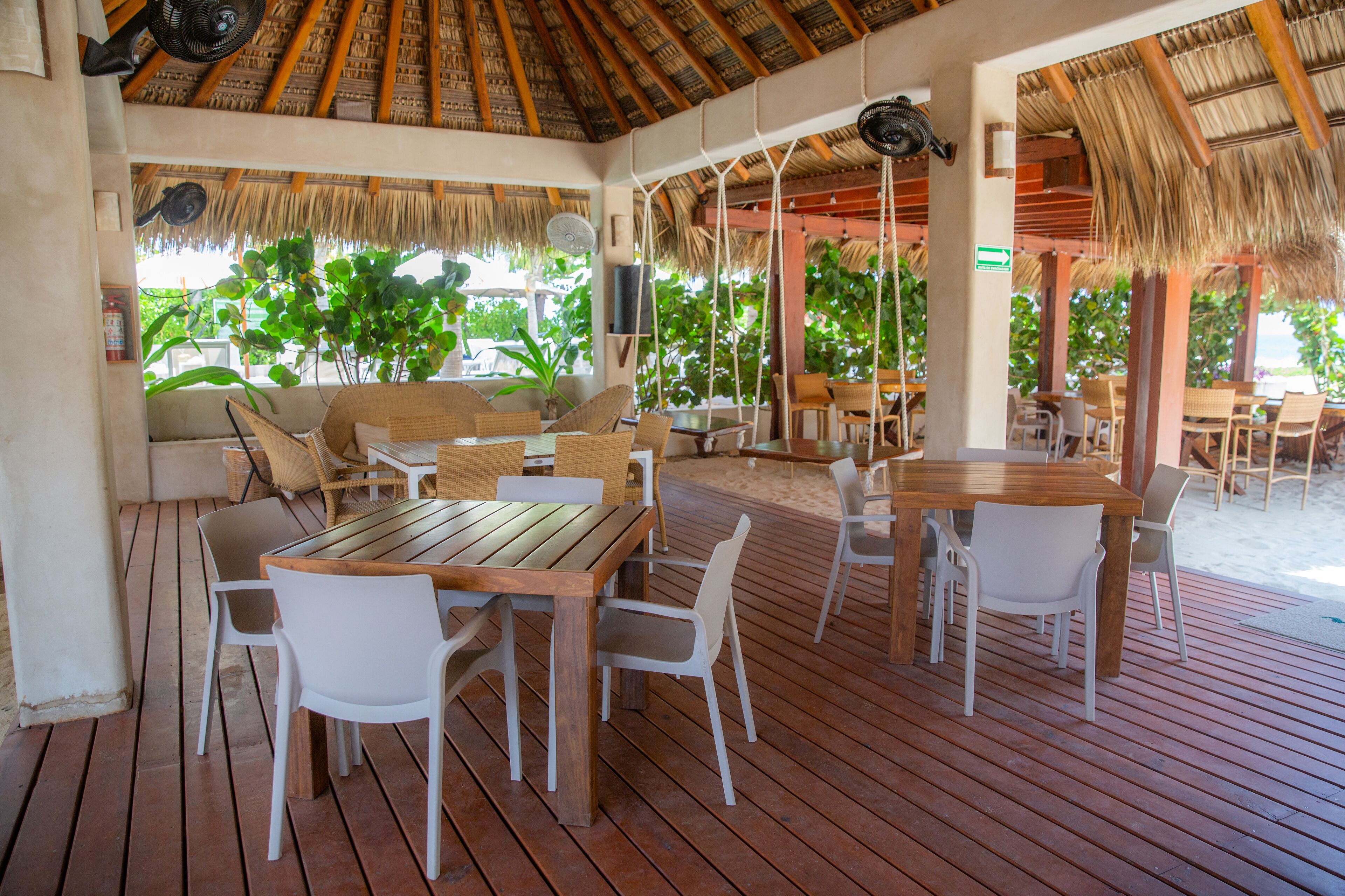 An outdoor seating area with wooden tables and white chairs, surrounded by thatched roof structures and lush greenery.