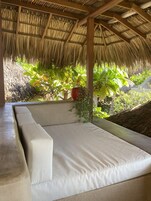 Un área de estar al aire libre con techo de paja, que incluye un banco acolchado, rodeada de abundante vegetación y plantas en macetas.