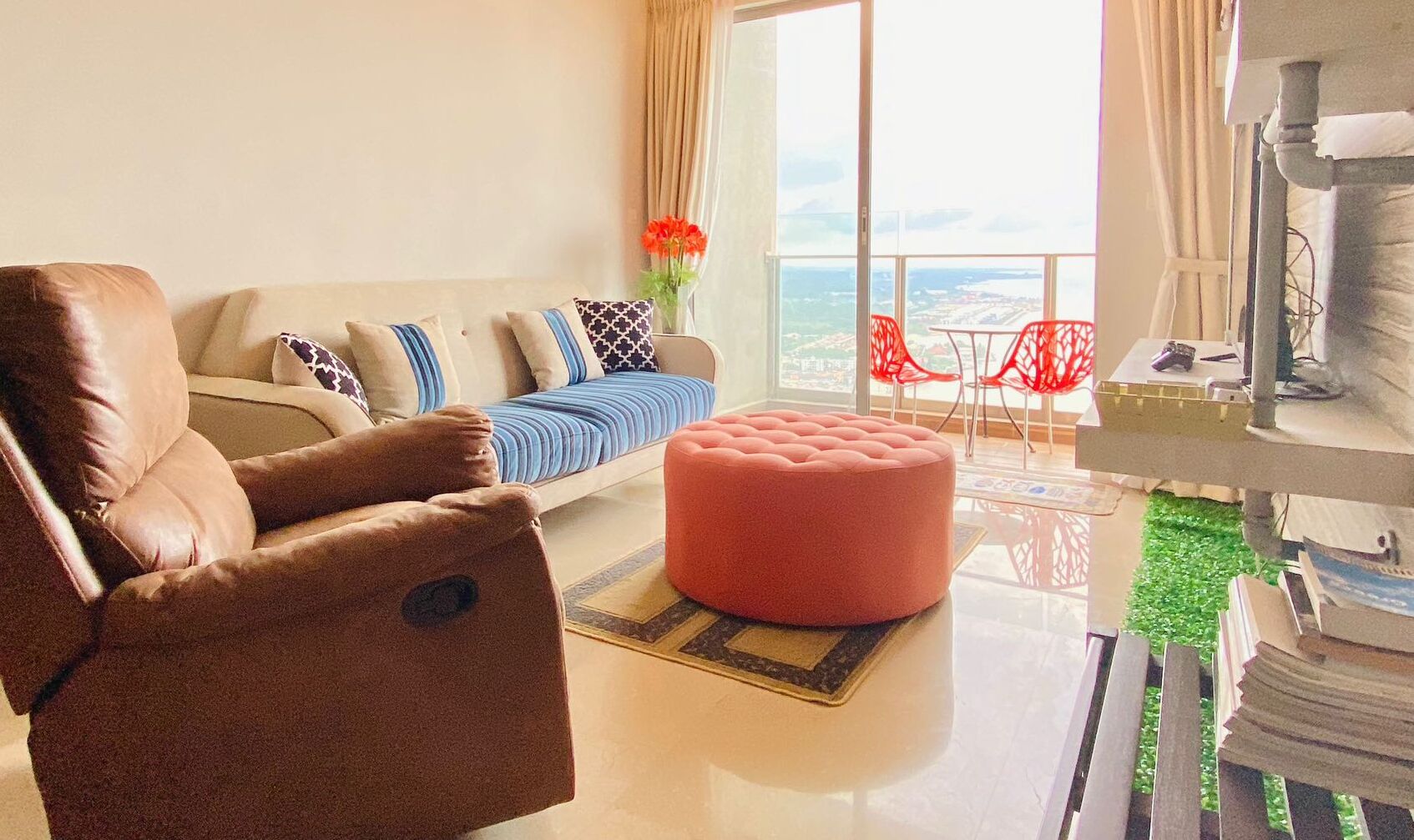 A modern living room with a leather sofa, a red ottoman, and a view of the city through large windows.
