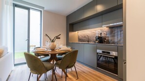 A modern kitchen with a built-in oven and a coffee machine. There is a dining area with a table set for four, and a large window providing natural light. - RUAH D - Delightful Studio by LovelyStay (Vila Nova de Gaia)