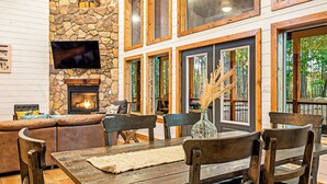 A cozy living room with a stone fireplace, comfortable seating, and large windows offering a view of the outdoors. - 2 Kings + Bunk Haus | Fun Games | Hot Tub | Wheelchair Access, Sleeps 11, Pets (Broken Bow)