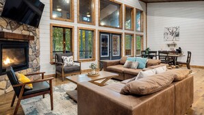 A spacious living room with a fireplace, large windows, and a stone accent wall. - 2 Kings + Bunk Haus | Fun Games | Hot Tub | Wheelchair Access, Sleeps 11, Pets (Broken Bow)