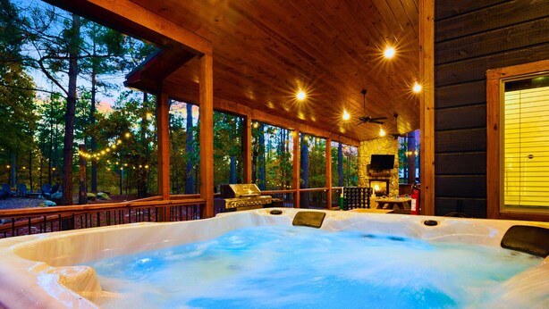 A hot tub in a wooden cabin with a stone wall, a grill, and a TV. - 2 Kings + Bunk Haus | Fun Games | Hot Tub | Wheelchair Access, Sleeps 11, Pets (Broken Bow)