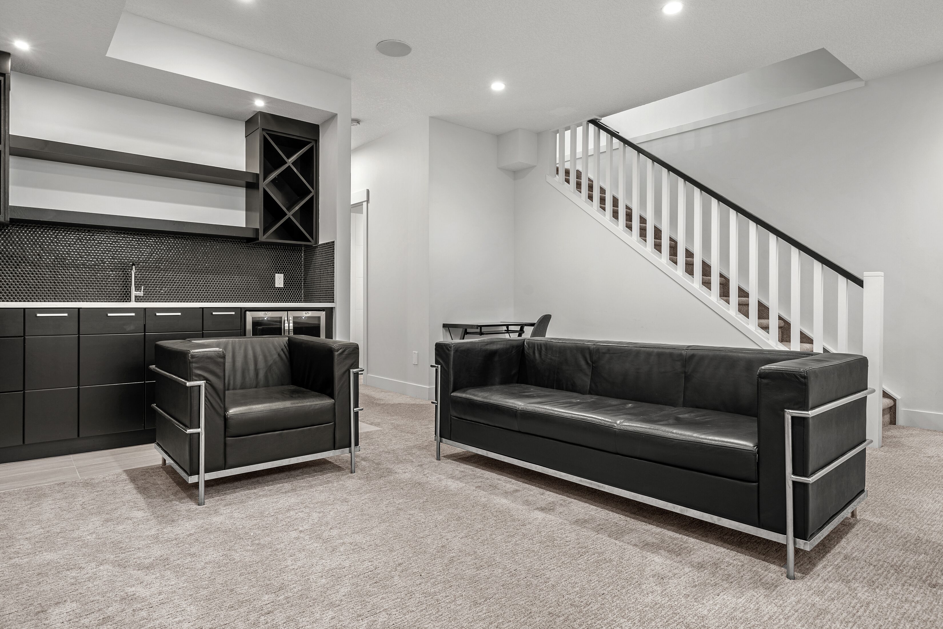 A modern living room with a black leather sofa and a matching armchair, a staircase, and a kitchen area with black cabinets and a tiled backsplash.