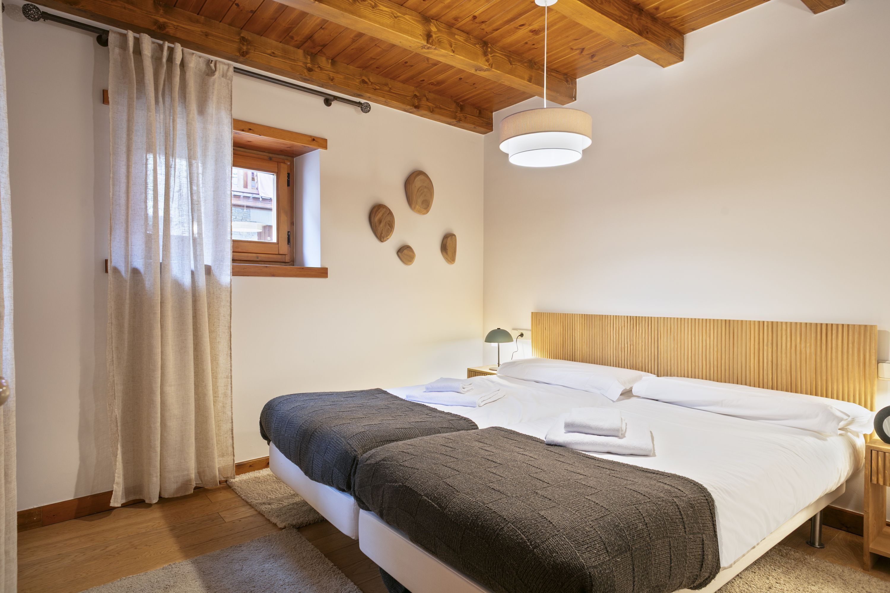 Un dormitorio con techo de madera, una cama con sábanas blancas, una mesita de noche con lámpara y una ventana con cortinas.