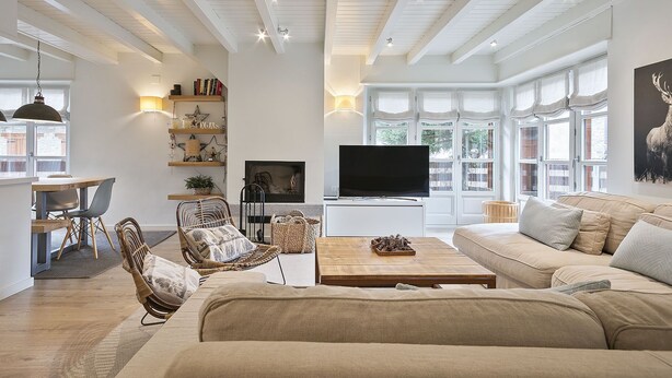 Ein geräumiges Wohnzimmer mit einer großen Couch, einem Couchtisch und einem Fernseher. Regale mit Dekorationen und ein Kamin sind ebenfalls vorhanden.