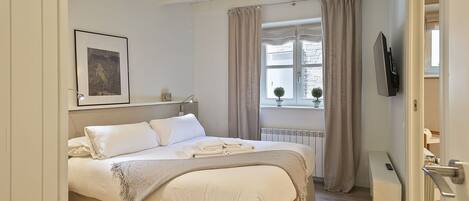 A bedroom with a bed, a bedside table, a TV mounted on the wall, and a window with curtains.