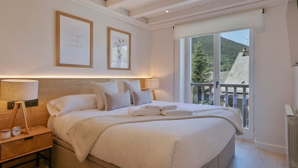 Un dormitorio con una cama grande, lámparas de noche, una mesita de noche, un cuadro enmarcado y una ventana con vistas a árboles y un edificio.