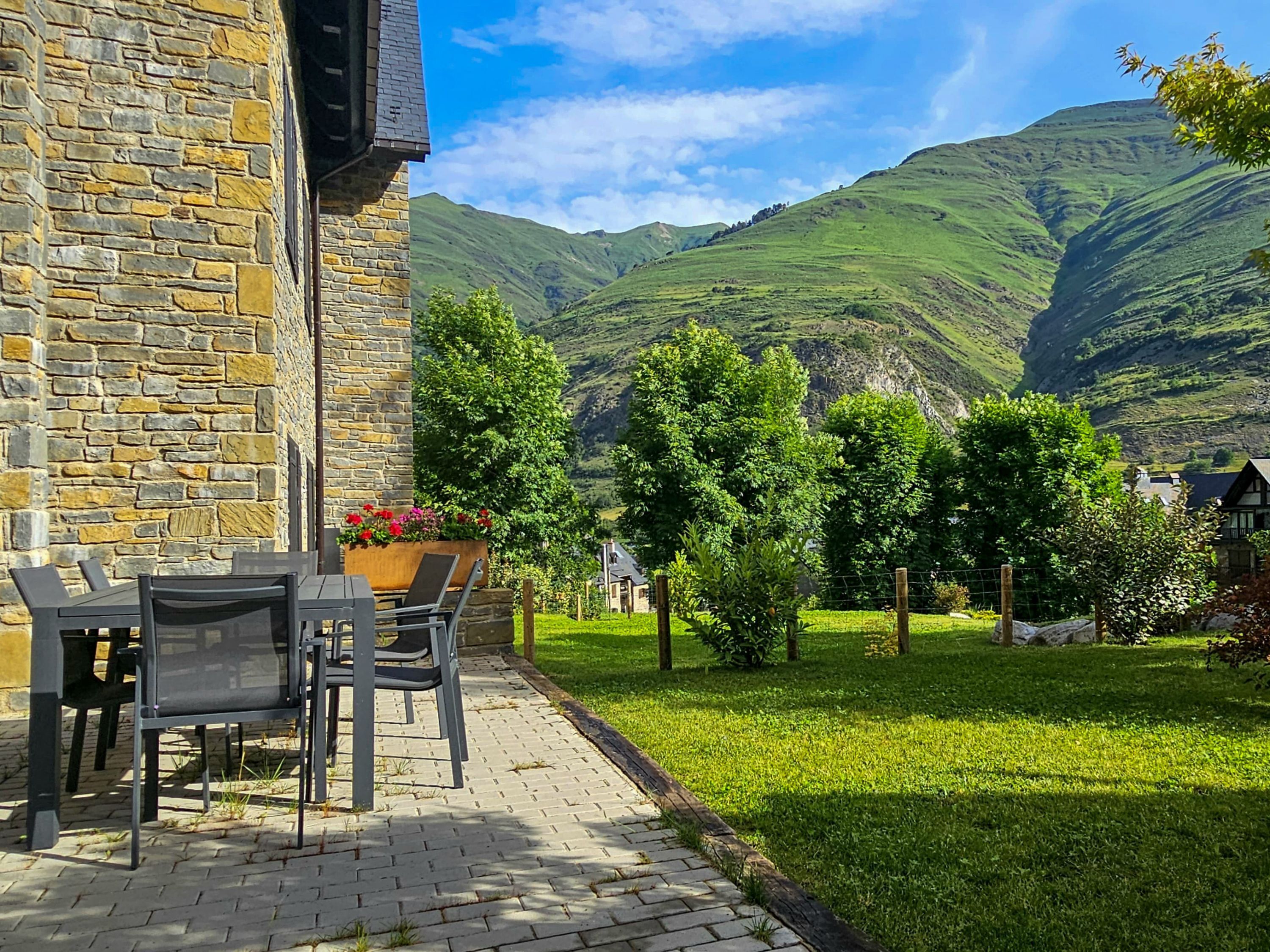 Edificio de piedra con una mesa y sillas en el patio, rodeado de un césped cuidado y árboles, con montañas al fondo.