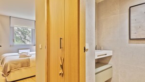 Een moderne hotelkamer met een houten garderobe, een bed met een beige plaid, een kleine badkamer met een wastafel en spiegel, en een raam met uitzicht op bomen.
