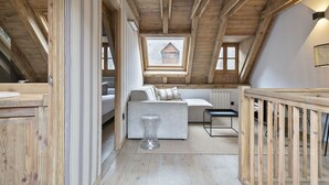 Et hyggeligt soveværelse på loftet med synlige træbjælker, en hemseng, et lille bord og udsigt til et hus gennem et ovenlys.