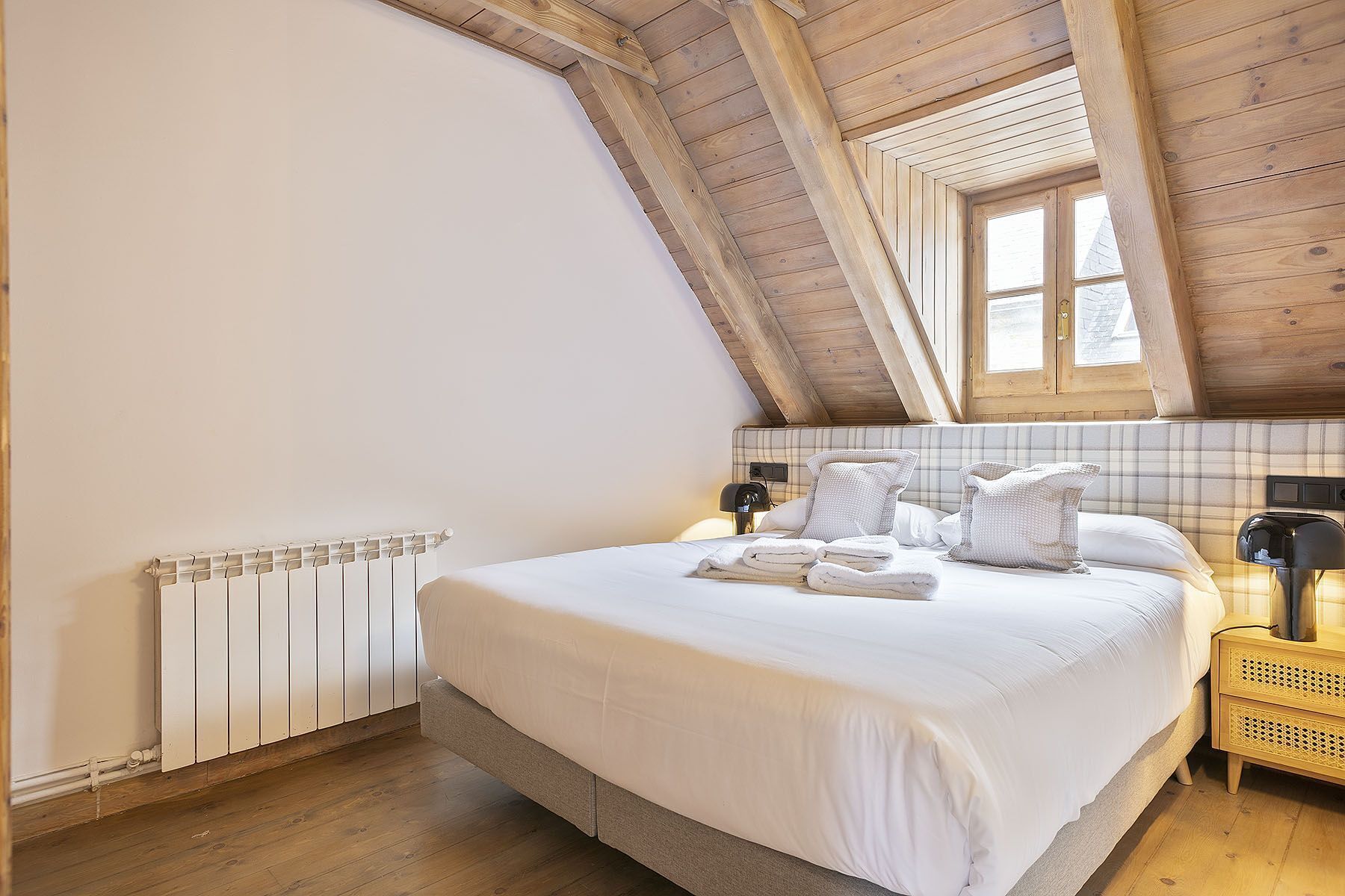 A bedroom with a sloped ceiling, a bed with white linens, a bedside table with a lamp, and a radiator.
