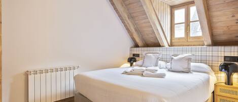 A bedroom with a sloped ceiling, a bed with white linens, a bedside table with a lamp, and a radiator.