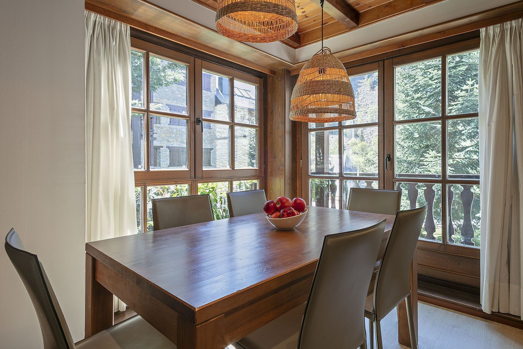 A dining area with a wooden table, four chairs, a bowl of fruit, and large windows with curtains.