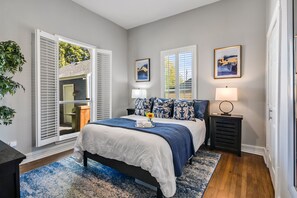 Un dormitorio con una cama, dos mesitas de noche, una mesita auxiliar, una ventana con persianas y una puerta.