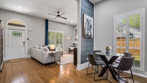 A modern living room with hardwood flooring, a white door, a ceiling fan, a gray sofa, a coffee table, and a dining area with a glass table and chairs. - Beautiful Retreat Near Historic Downtown McKinney (McKinney)