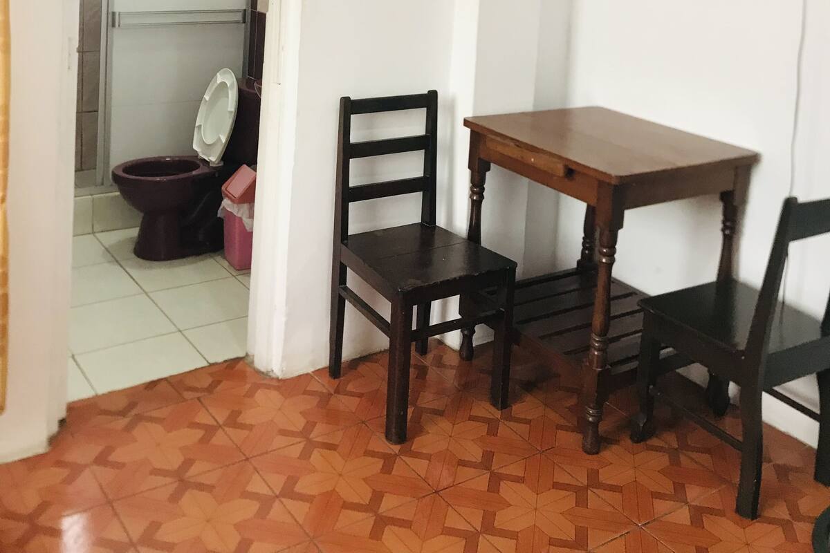 Ein kleiner Raum mit gefliestem Boden, einem Holztisch, zwei Stühlen, einem an der Wand befestigten Fernseher und einer Toilette, die durch eine offene Tür sichtbar ist.