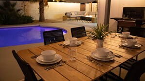 A dining table set for six with white plates, bowls, and cutlery, surrounded by chairs, with a pool and outdoor seating area in the background.