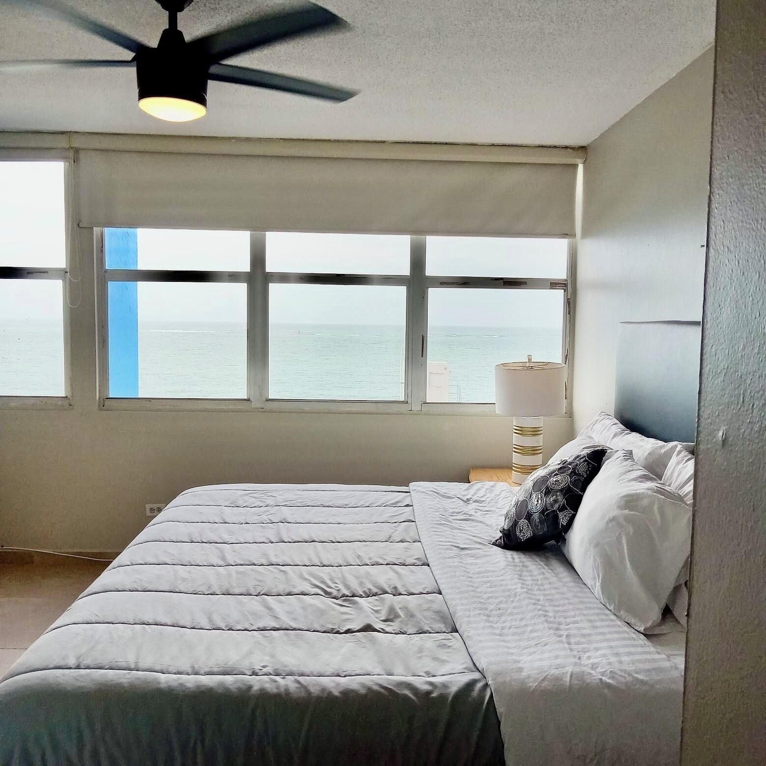Une chambre à coucher avec un lit, un ventilateur de plafond, une fenêtre donnant sur la mer et une lampe de chevet.