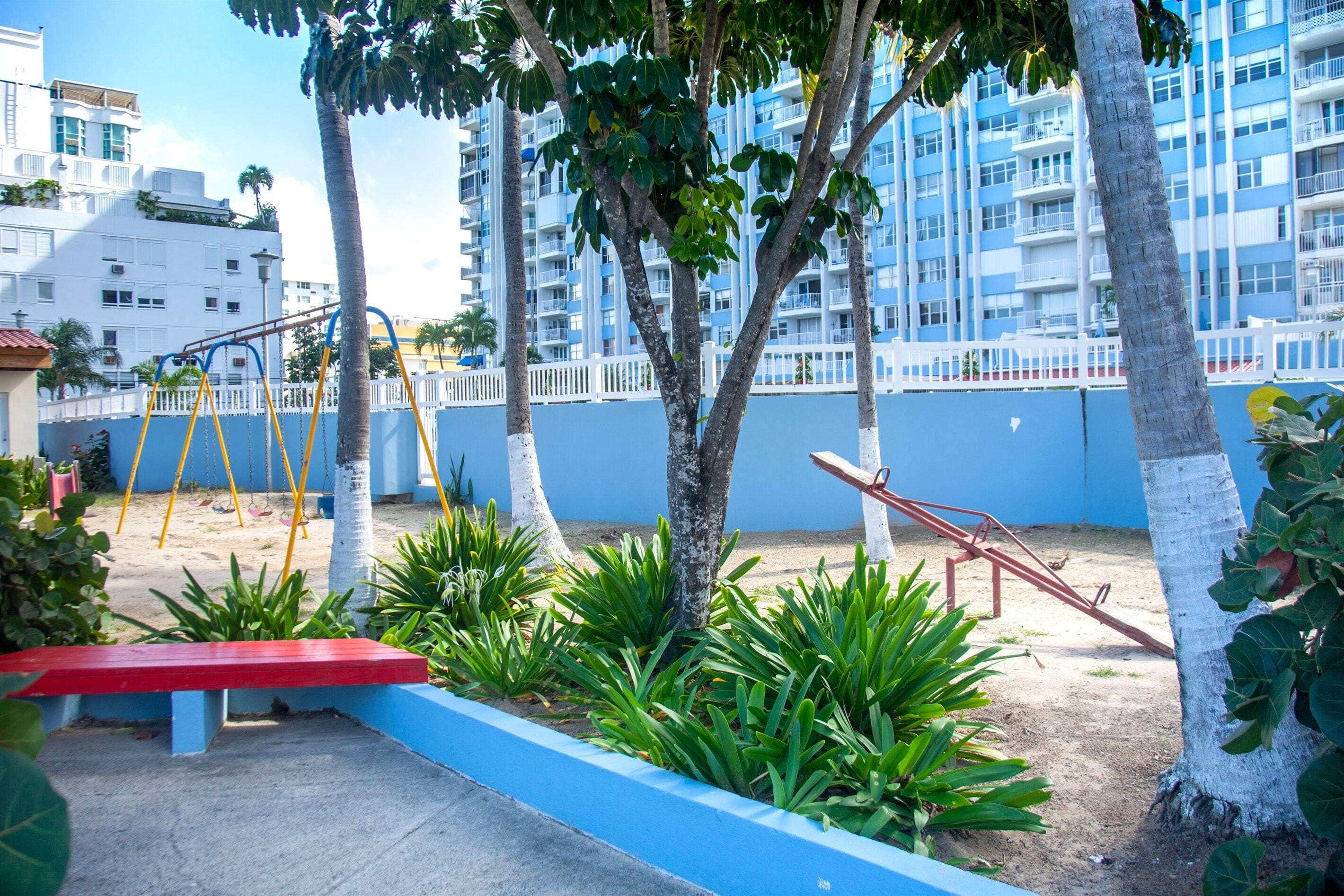 A playground with swings and a slide, surrounded by palm trees and a blue wall.