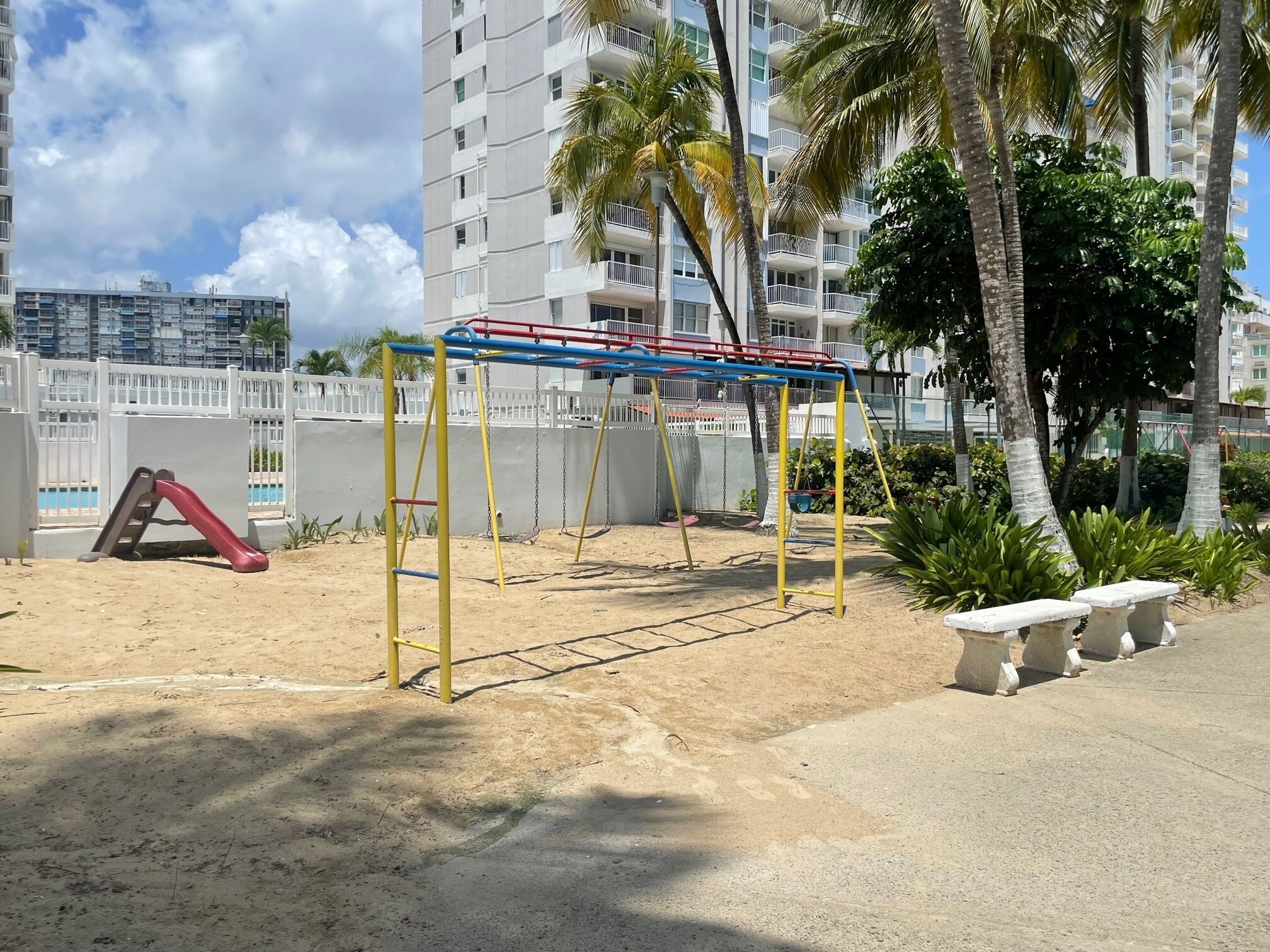 A playground with a slide and swings, surrounded by palm trees and a white fence.