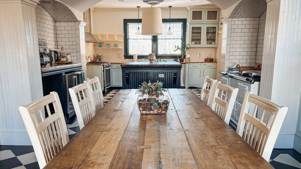 Ein langer Holztisch mit weißen Stühlen, eine zentrale Blumenkomposition und eine Küche mit gefliestem Rücksprung und Holzschränken.