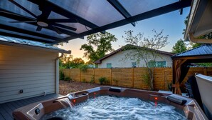 Un bain à remous doté d’une terrasse en bois et d’un espace abrité, entouré d’une clôture en bois et avec un bâtiment en briques en arrière-plan.