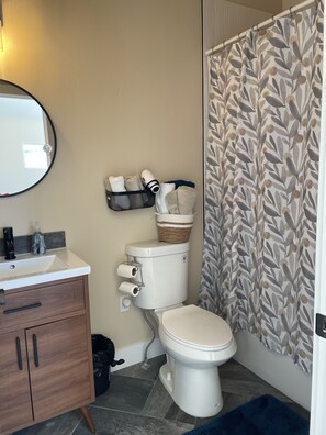 A bathroom with a toilet, a sink, and a mirror. There is a basket on the toilet tank and a towel rack on the wall.