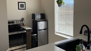 Una cocina moderna con horno integrado, microondas y refrigerador, un ventanal con persianas y una planta colgante.