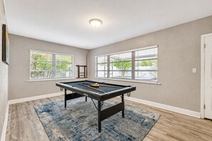 Mesa de bilhar em sala com janelas amplas e luminária no teto.