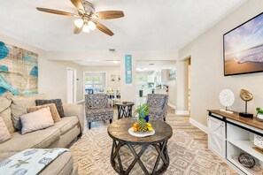 A spacious living room with a ceiling fan, a coffee table with fruit, patterned chairs, and a kitchen in the background.