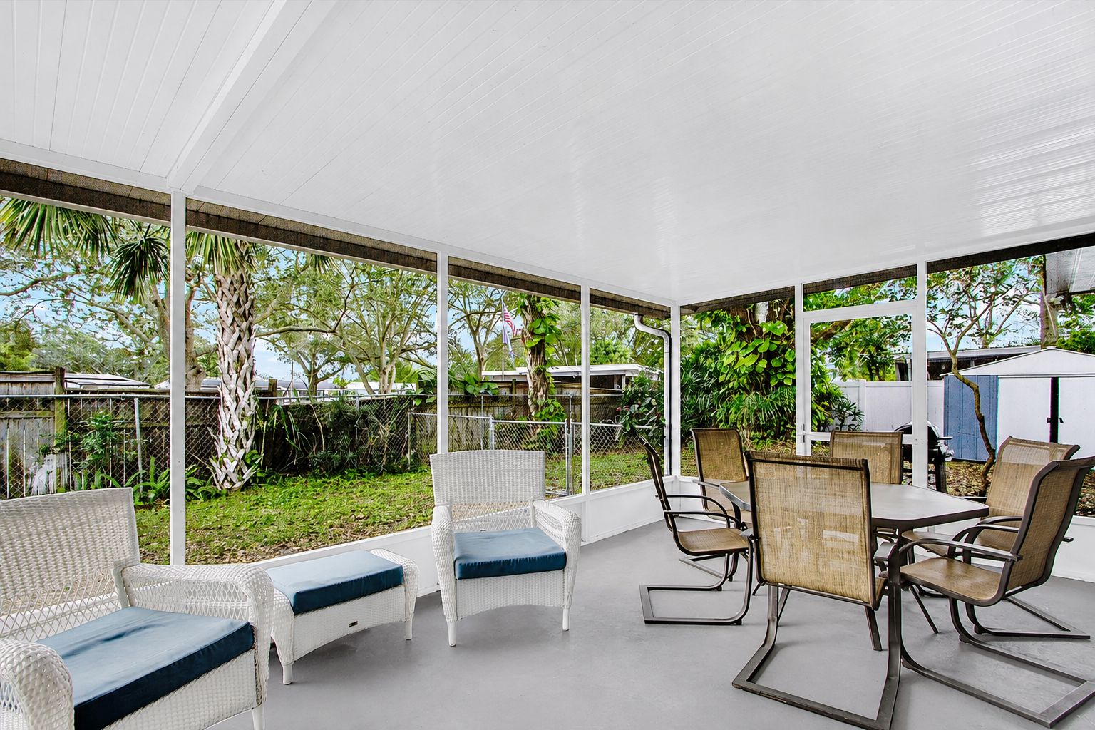 Área de varanda coberta com móveis de vime e uma mesa, cercada por um muro branco e vegetação.