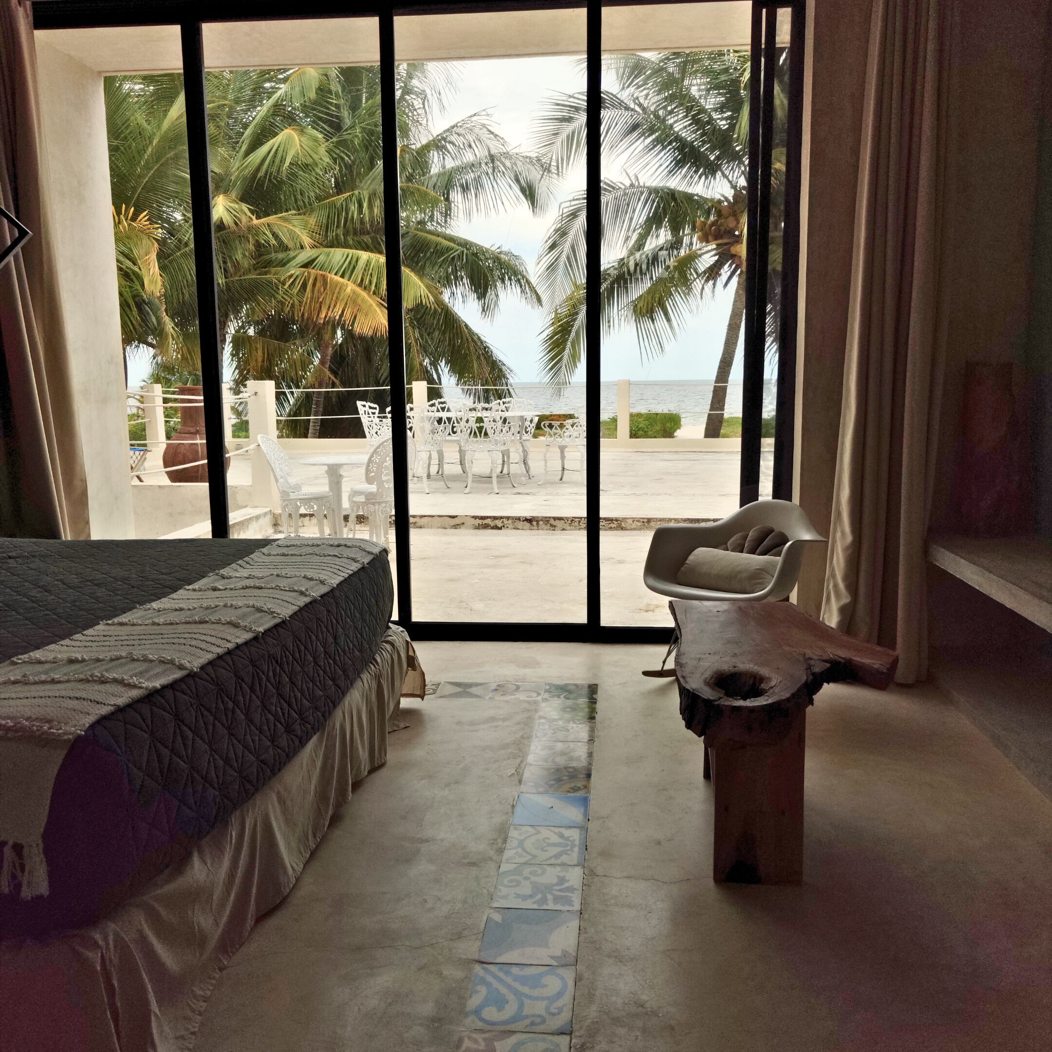 A room with a bed, a small table, and a view of the beach and palm trees through large glass doors.