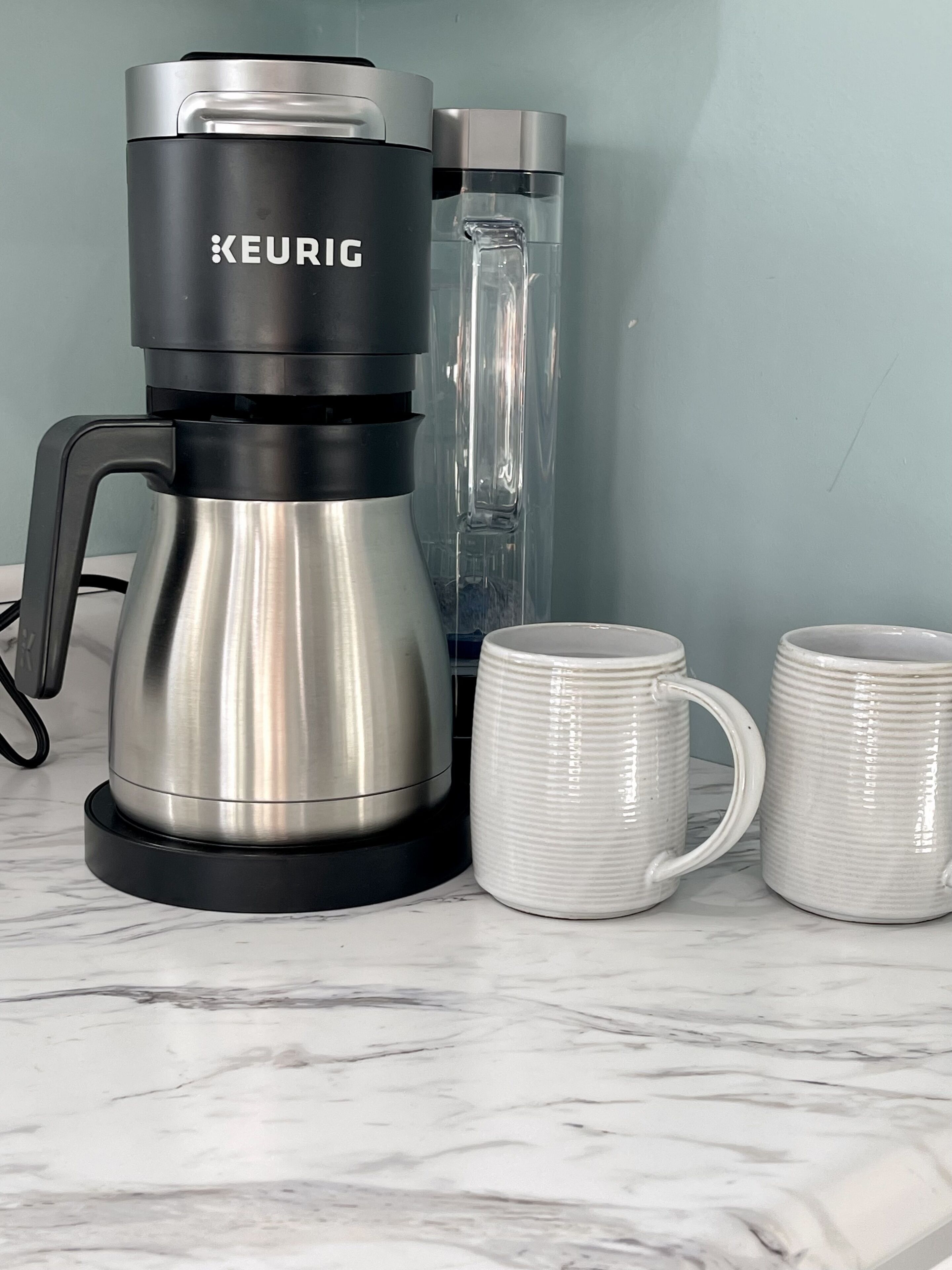 A Keurig coffee maker on a marble countertop with two white mugs beside it.