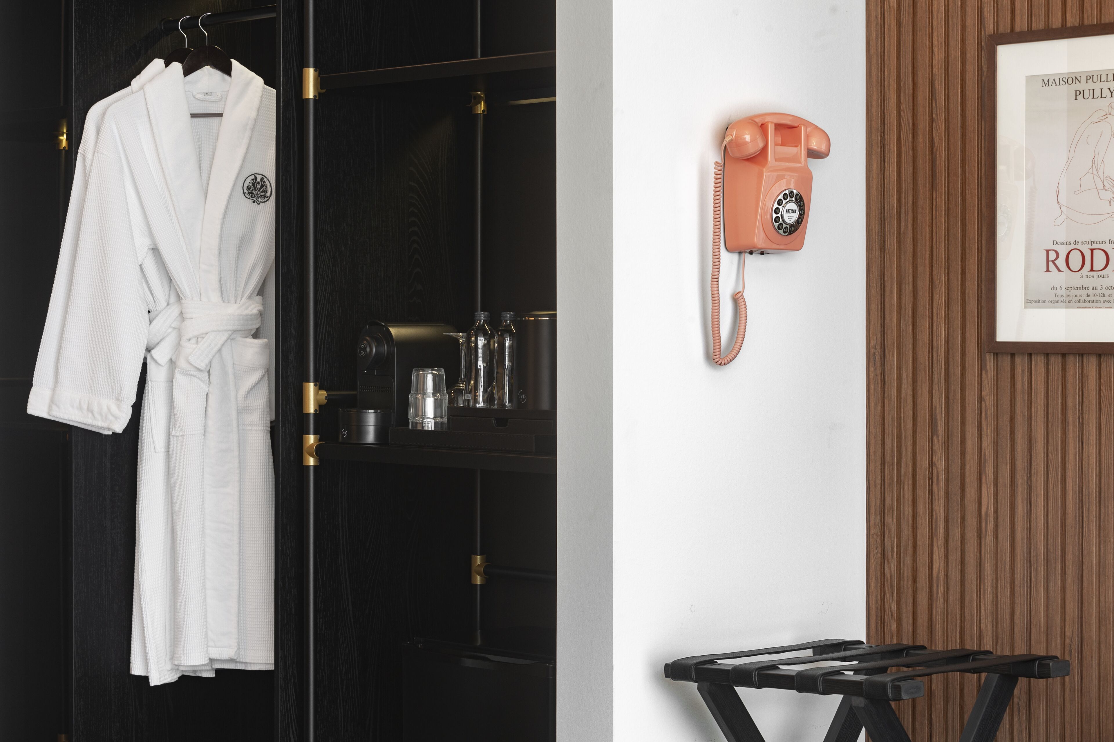 A hotel room with a dark wooden wardrobe, a white robe hanging on the door, a vintage orange telephone, and a framed map on the wall.
