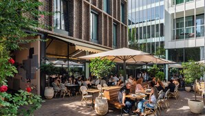 Ein Straßencafé mit Tischen und Stühlen, umgeben von Pflanzkübeln und einer Ziegelwand.