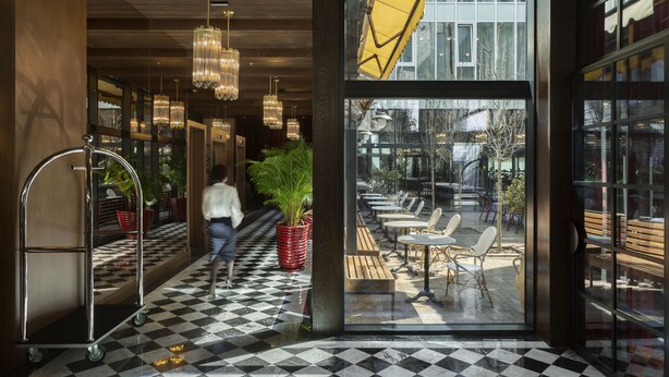 Eine Hotelhalle mit Schachbrettmuster-Boden, einer großen Glasdoor zu einem Außenbereich mit Sitzgelegenheiten und einem Wagen in der Nähe des Eingangs.