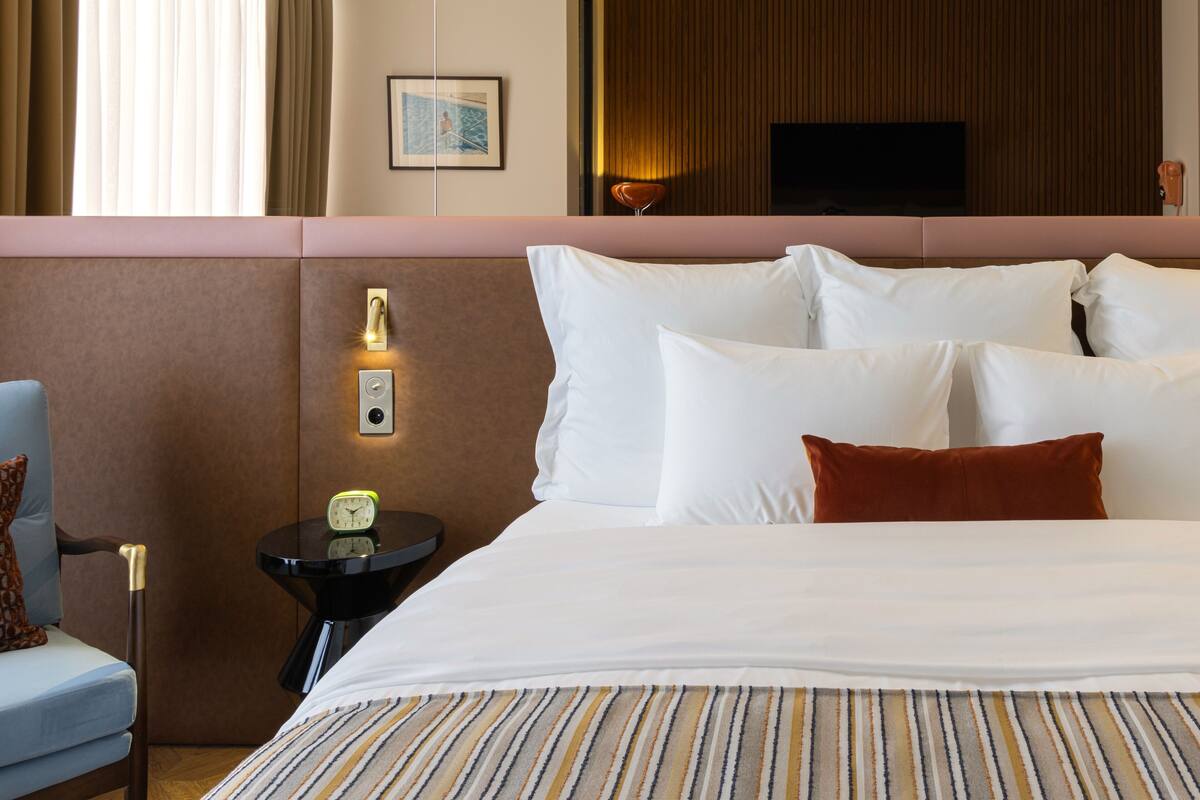 A hotel room with a large bed, a bedside table with a clock, a chair, and a television on the wall.