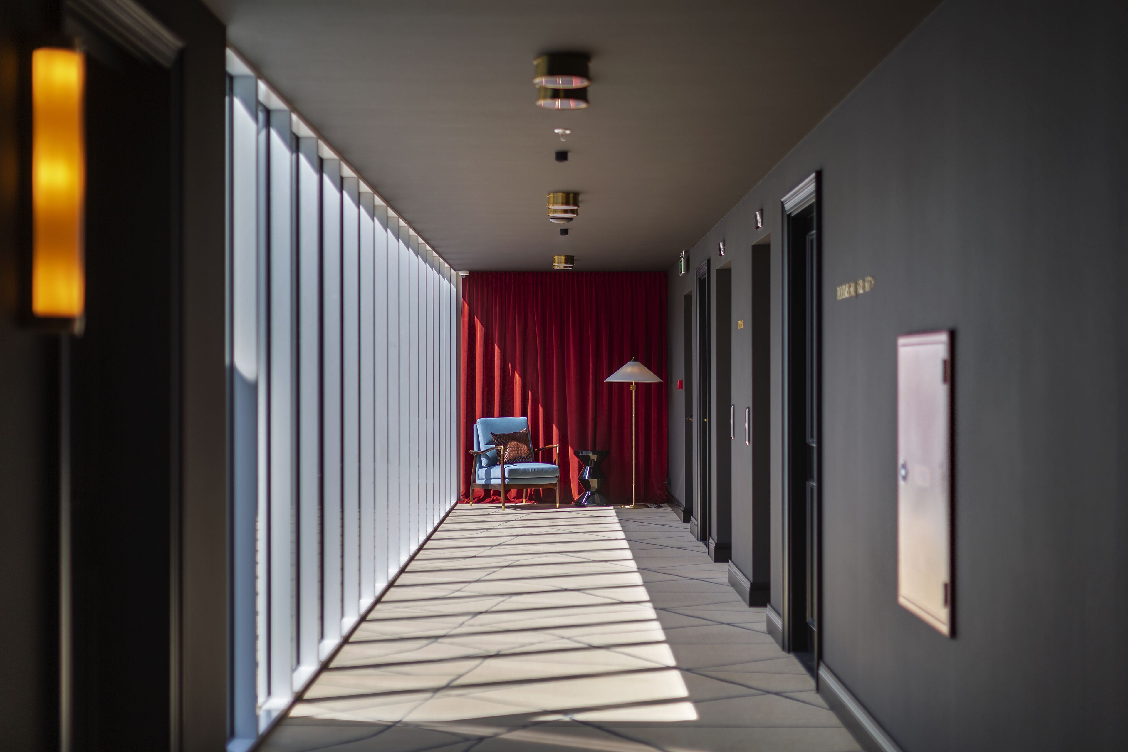 Un couloir avec un rideau rouge, une chaise bleue et un lampadaire.