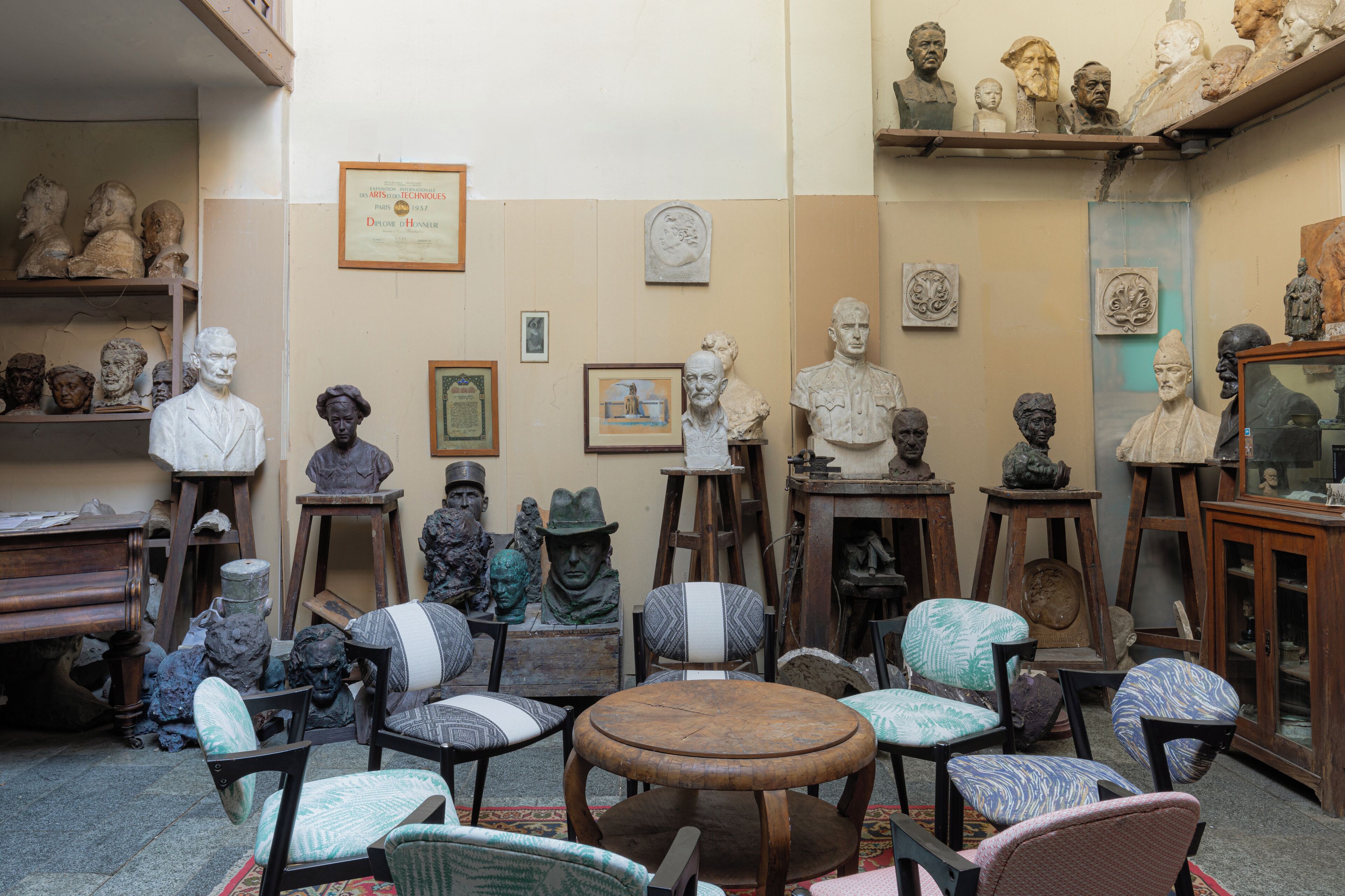 A room with various sculptures and busts displayed on shelves and tables, surrounded by upholstered chairs and a round wooden table.