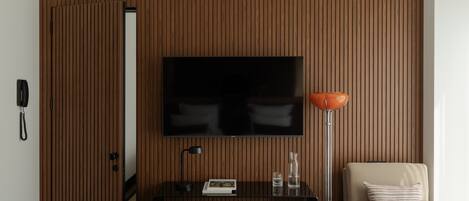 Quarto de hotel moderno com parede revestida de madeira, televisão de tela plana fixada acima de uma mesa, uma cadeira, abajur e uma cama com travesseiro listrado.