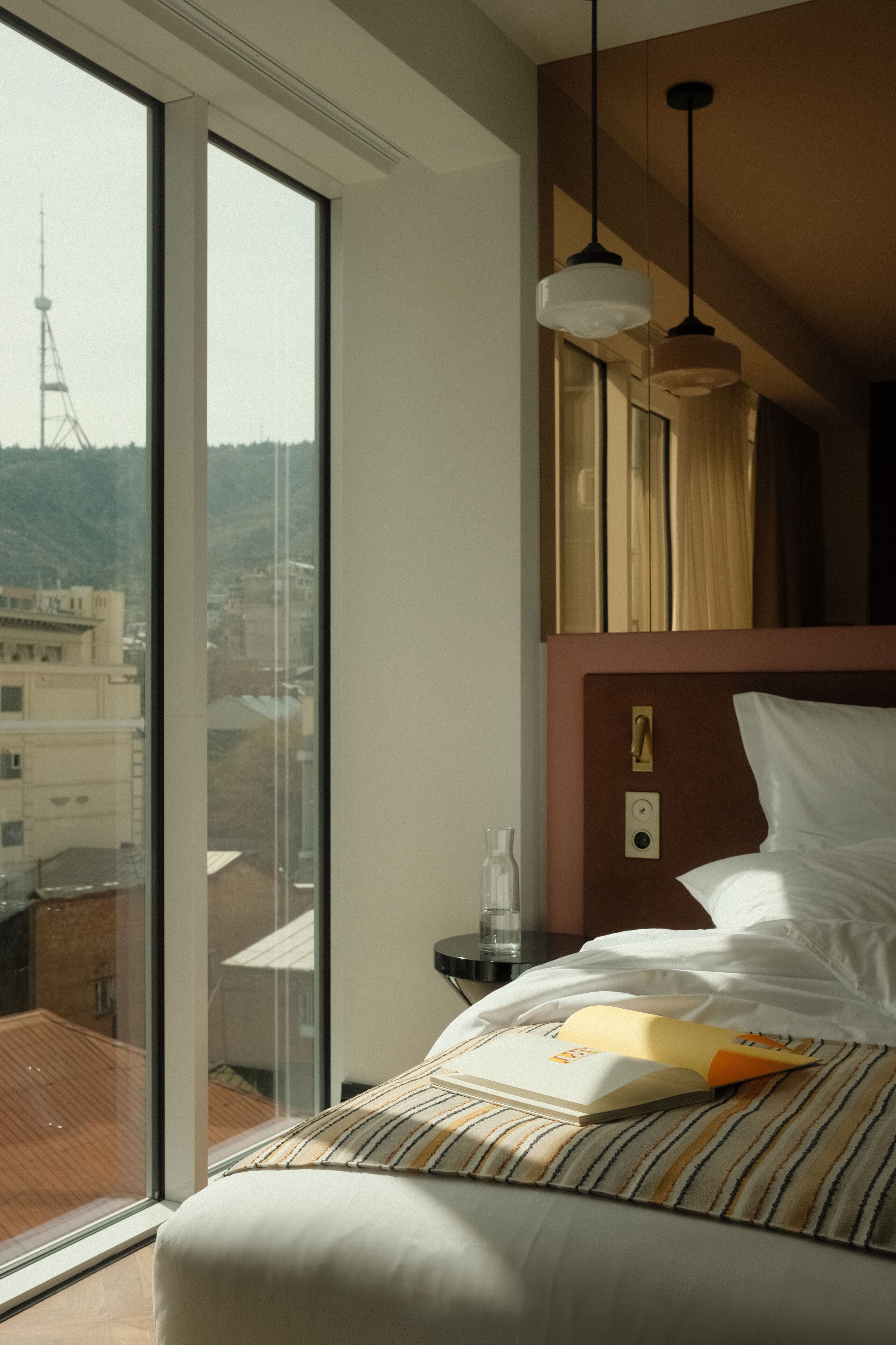 Une chambre d’hôtel avec une grande fenêtre, un lit avec un oreiller à rayures et un luminaire suspendu.