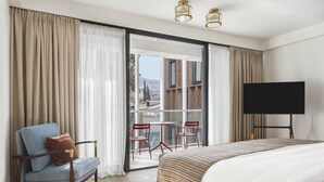 Ein Hotelzimmer mit Bett, Fernseher, Sessel, Tisch und Blick auf Gebäude durch das Fenster.
