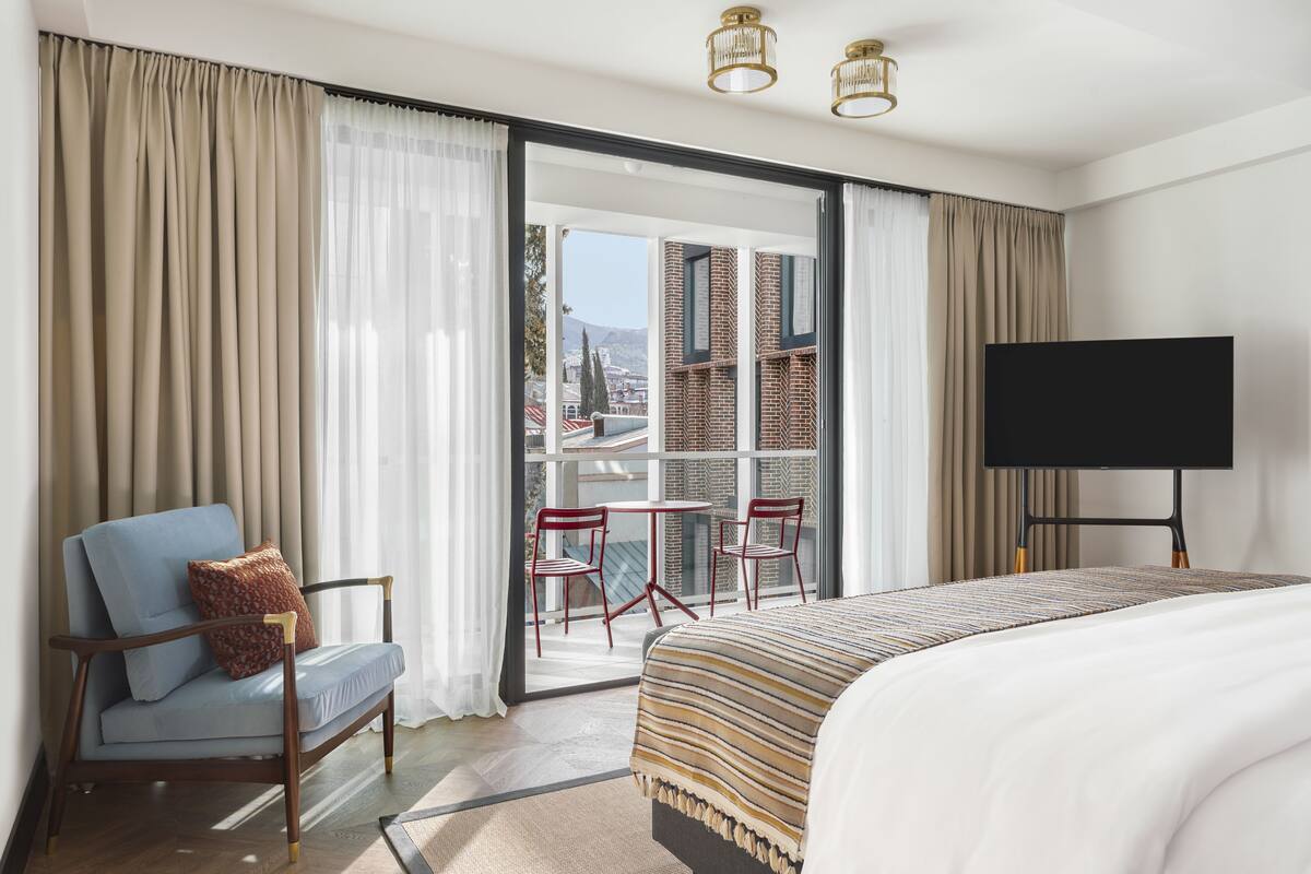 A hotel room with a bed, a TV, a chair, a table, and a view of buildings through the window.