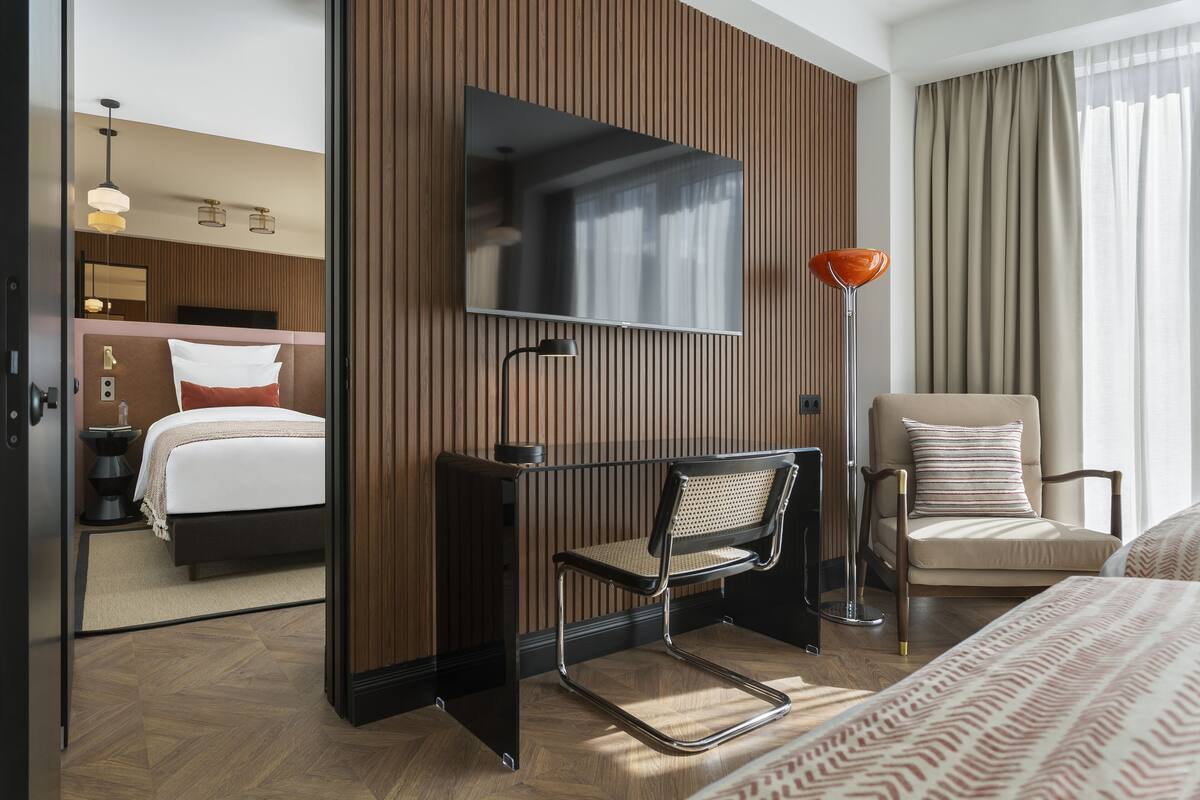 A modern hotel room with a large bed, a desk with a chair, a television, and a chair with a cushion.