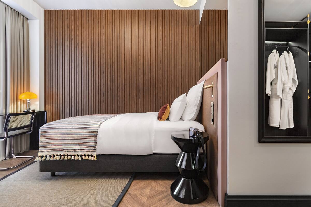 A modern hotel room with a large bed, a wooden panel wall, a black coffee maker, and a coat rack with white robes.