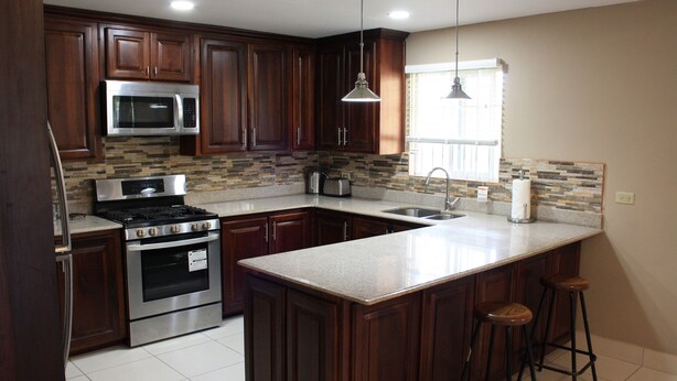 A kitchen with dark wood cabinets, stainless steel appliances, a granite countertop, and a window with blinds. - Soleil Hartland Palms (St. Ann's Bay)