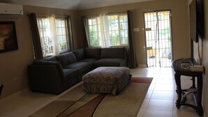 A living room with a sectional sofa, a patterned ottoman, a small table, and a TV. - Soleil Hartland Palms (St. Ann's Bay)