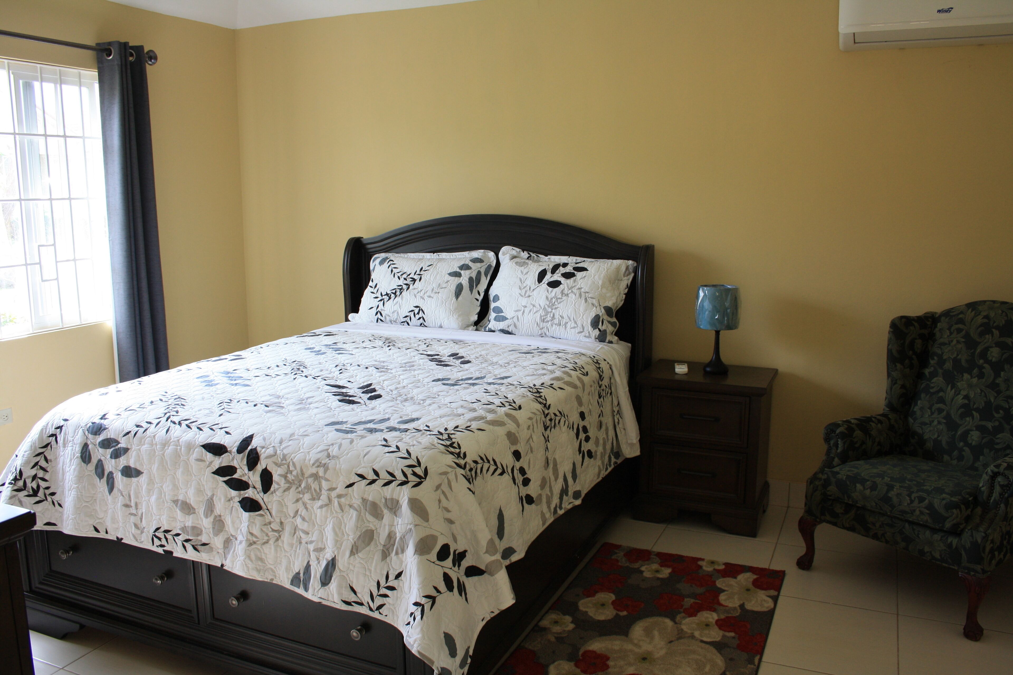 Un dormitorio con cama, mesita de noche, silla y ventana con cortinas.